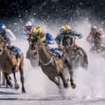 Several jockeys racing their horses at full gallop in the snow on an outdoor horse racing track.