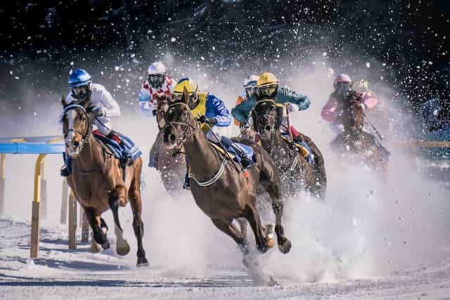 Several jockeys racing their horses at full gallop in the snow on an outdoor horse racing track.