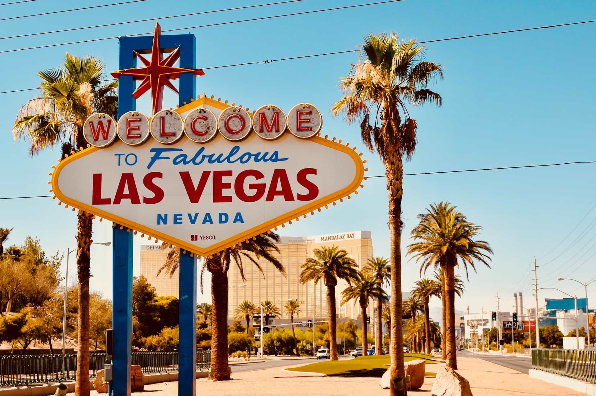 The iconic ‘Welcome to Las Vegas’ entrance sign in the city center, surrounded by palm trees and the Mandalay Bay hotel and casino standing behind it.