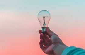 A person’s outstretched hand holding up a lightbulb against a blue and orange background.