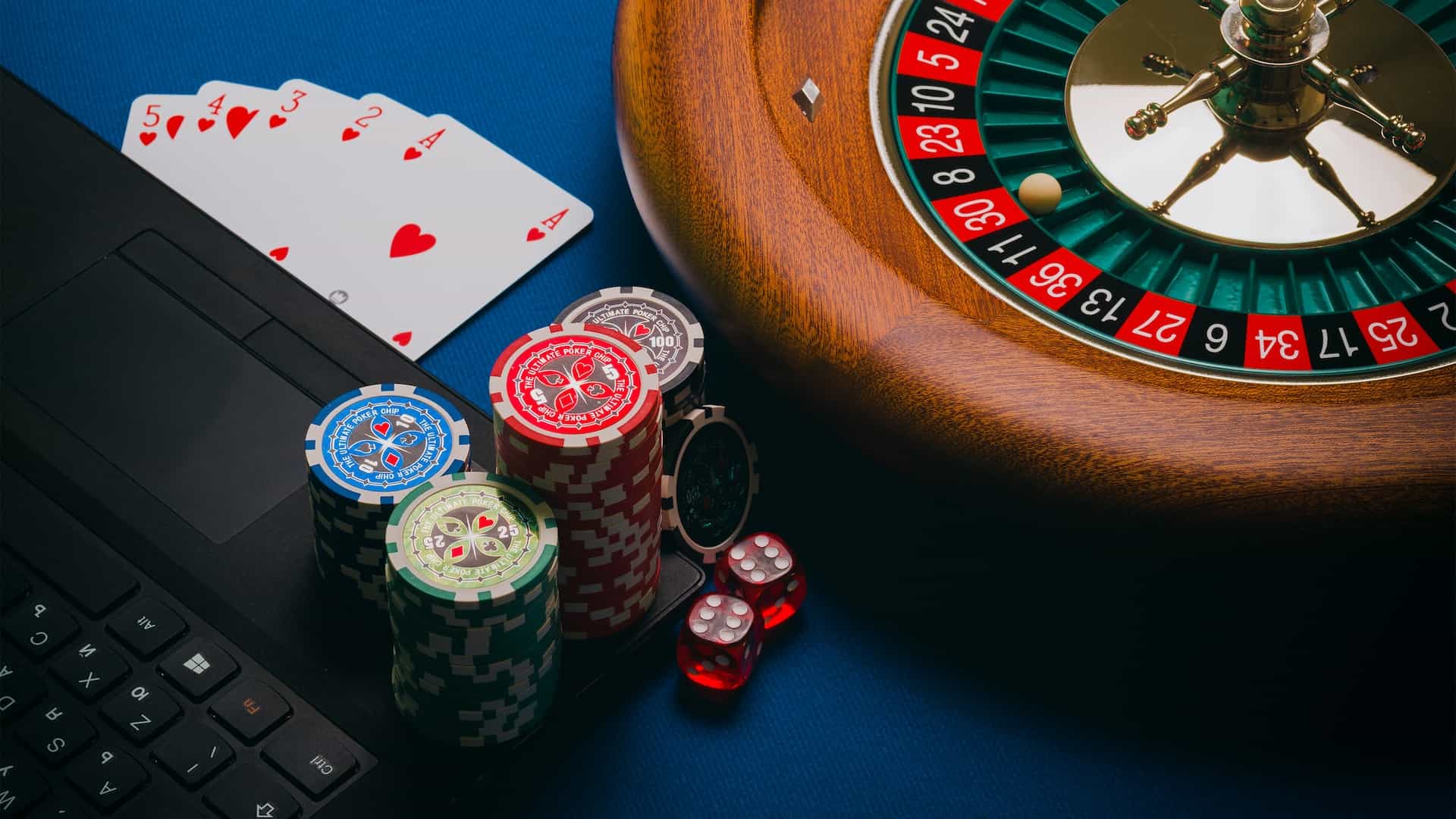 The corner of a laptop with several stacks of poker chips placed on it, placed next to a hand of playing cards and a roulette table. 