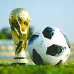 The golden World Cup trophy sits on a soccer field next to a soccer ball.