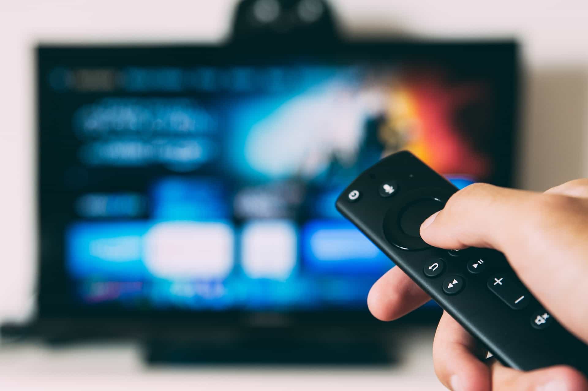 A person’s hand holding a smart TV remote which is being pointed at a television in the background.