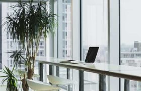 A laptop sitting at an empty workspace in a corporate office facing large floor to ceiling windows.