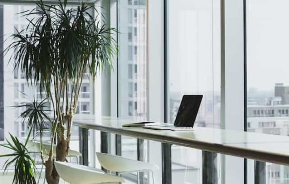 A laptop sitting at an empty workspace in a corporate office facing large floor to ceiling windows.