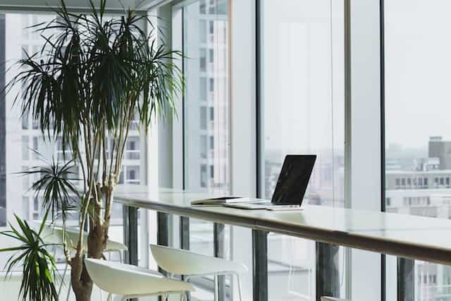A laptop sitting at an empty workspace in a corporate office facing large floor to ceiling windows.