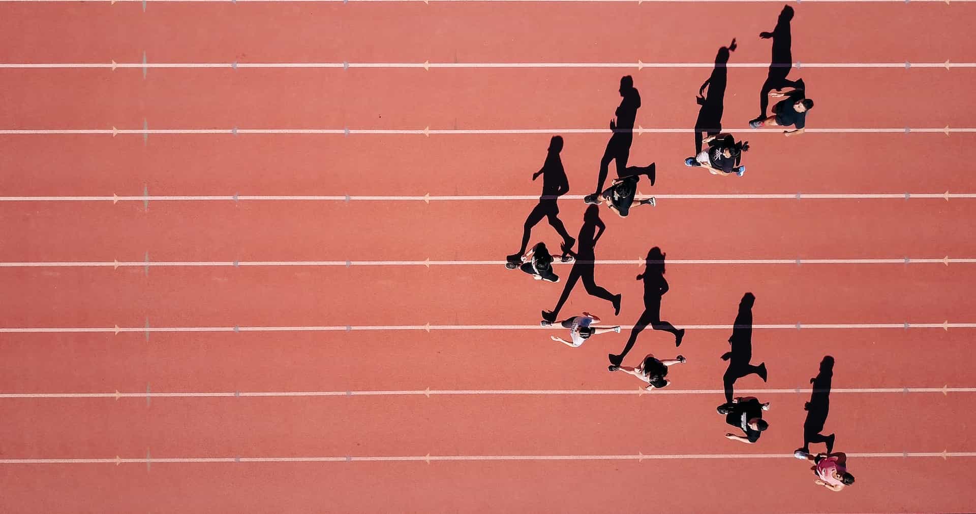 A top down view of several athlete runners training on an outdoor track and field course, with each runner’s shadow being dramatically cast on the ground thanks to the sun’s position.
