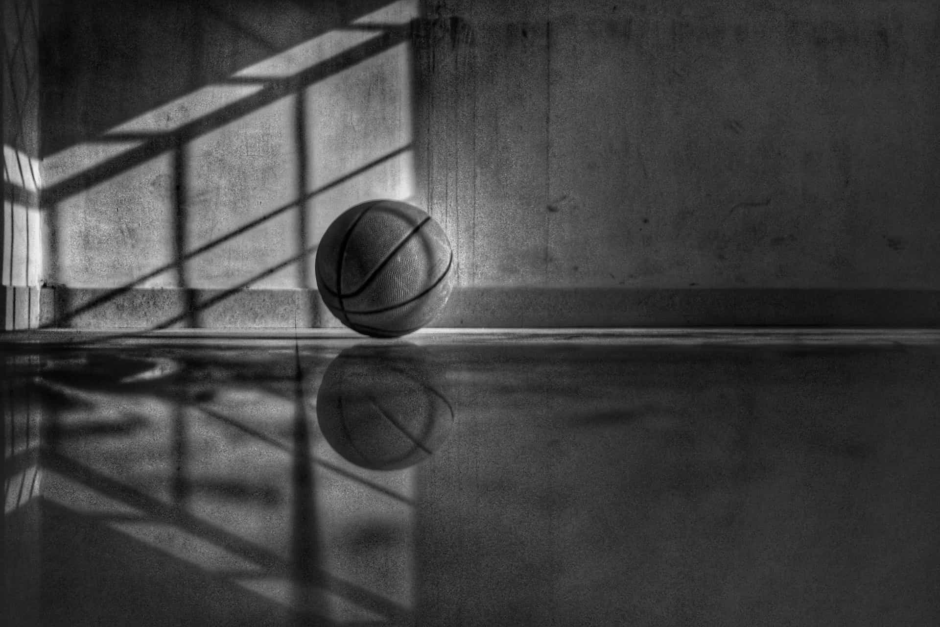 A lonely basketball sitting on a court in the corner near a wall with the outdoor light shining on it.