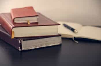 Three heavy books stacked on top one another on a desk next to an open notebook with a pen on top.