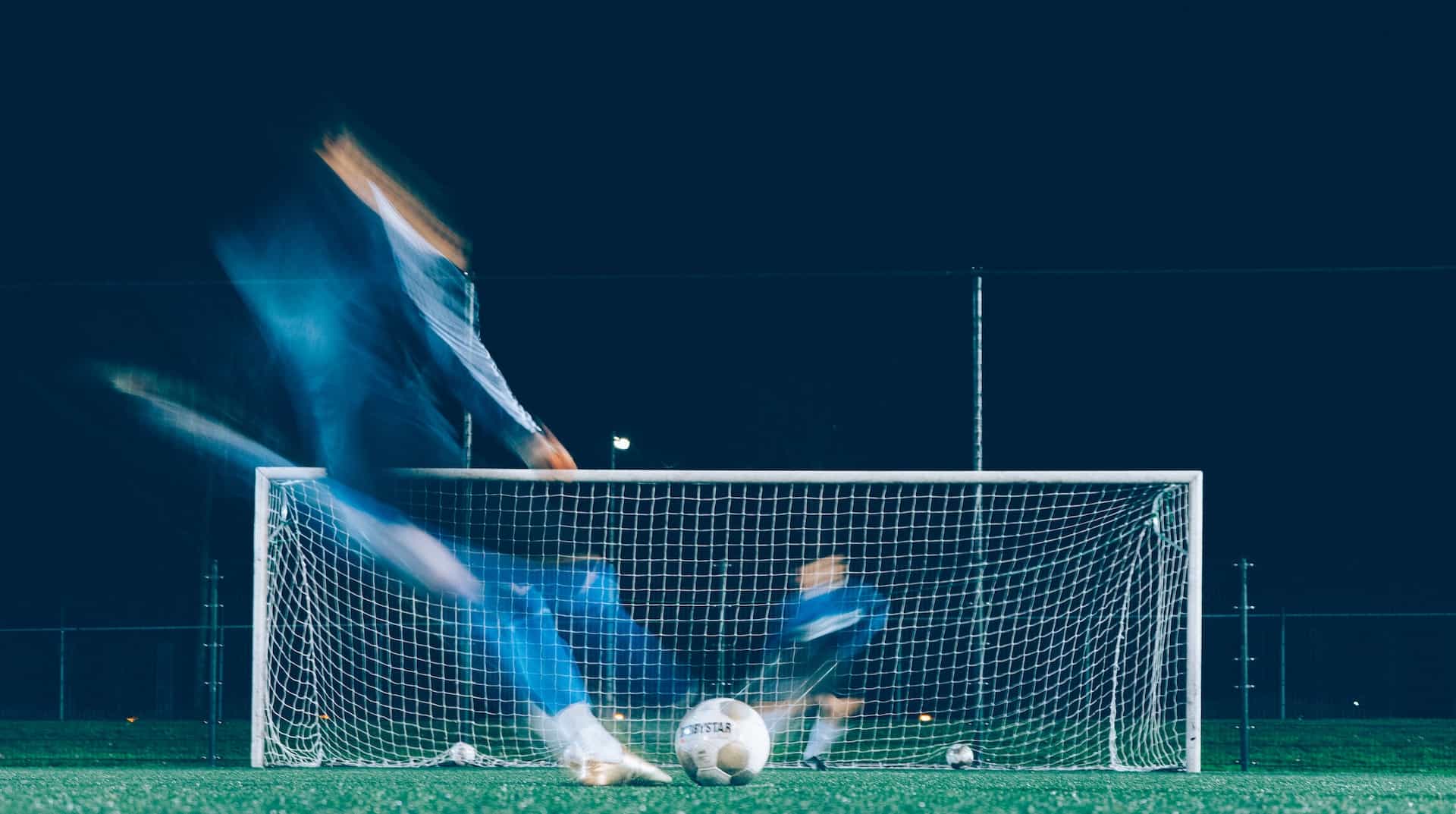 Blurred image of soccer players kicking a ball into a net.