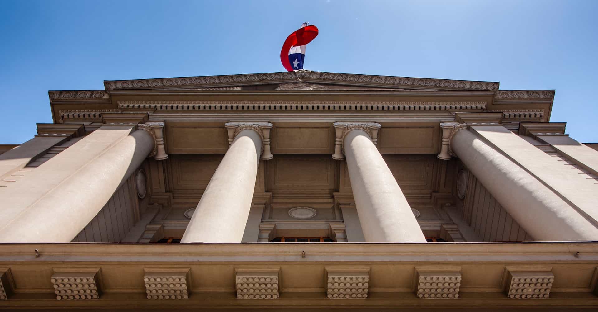 From below, a large building with columns and the Chilean flag waving at its front, in Santiago, Chile.