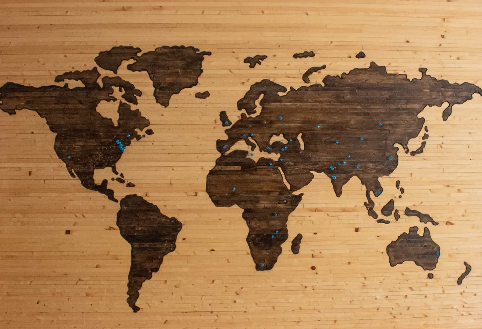 A brown wooden map of the world with blue pins marking locations.