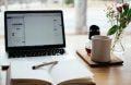 An open notebook lying flat on a table in front of a laptop next to a coffee mug and flower vase.