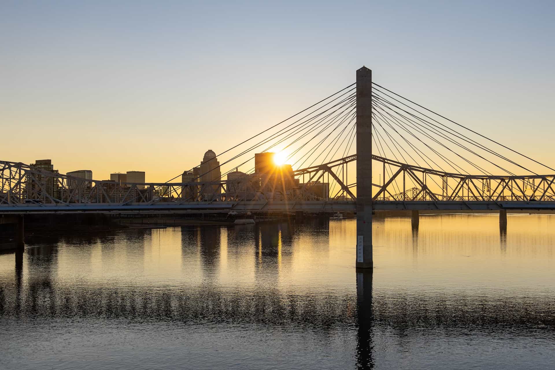 A large bridge cutting across the Ohio River in Louisville, Kentucky, with the city’s tall buildings seen off in the distance behind the bridge and water.