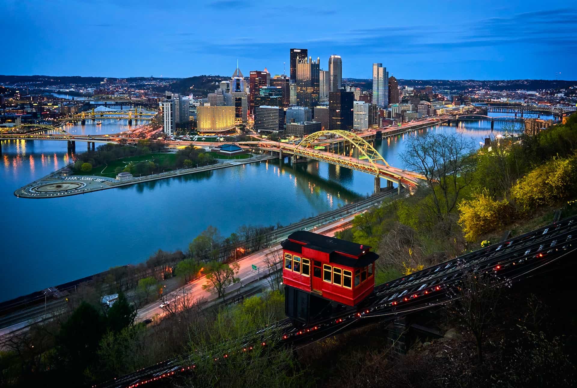 The city of Pittsburgh, Pennsylvania, with the dense downtown district seen surrounded by a large river and several bridges cutting across it.