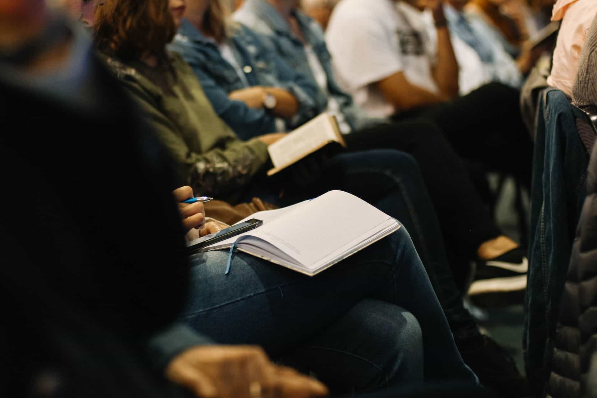 A group of people in an audience sit with notebooks and pens.
