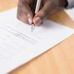 A person’s hand holding a pen and signing a written agreement or contract at the bottom of the page.