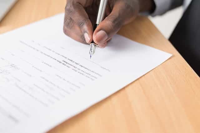 A person’s hand holding a pen and signing a written agreement or contract at the bottom of the page.