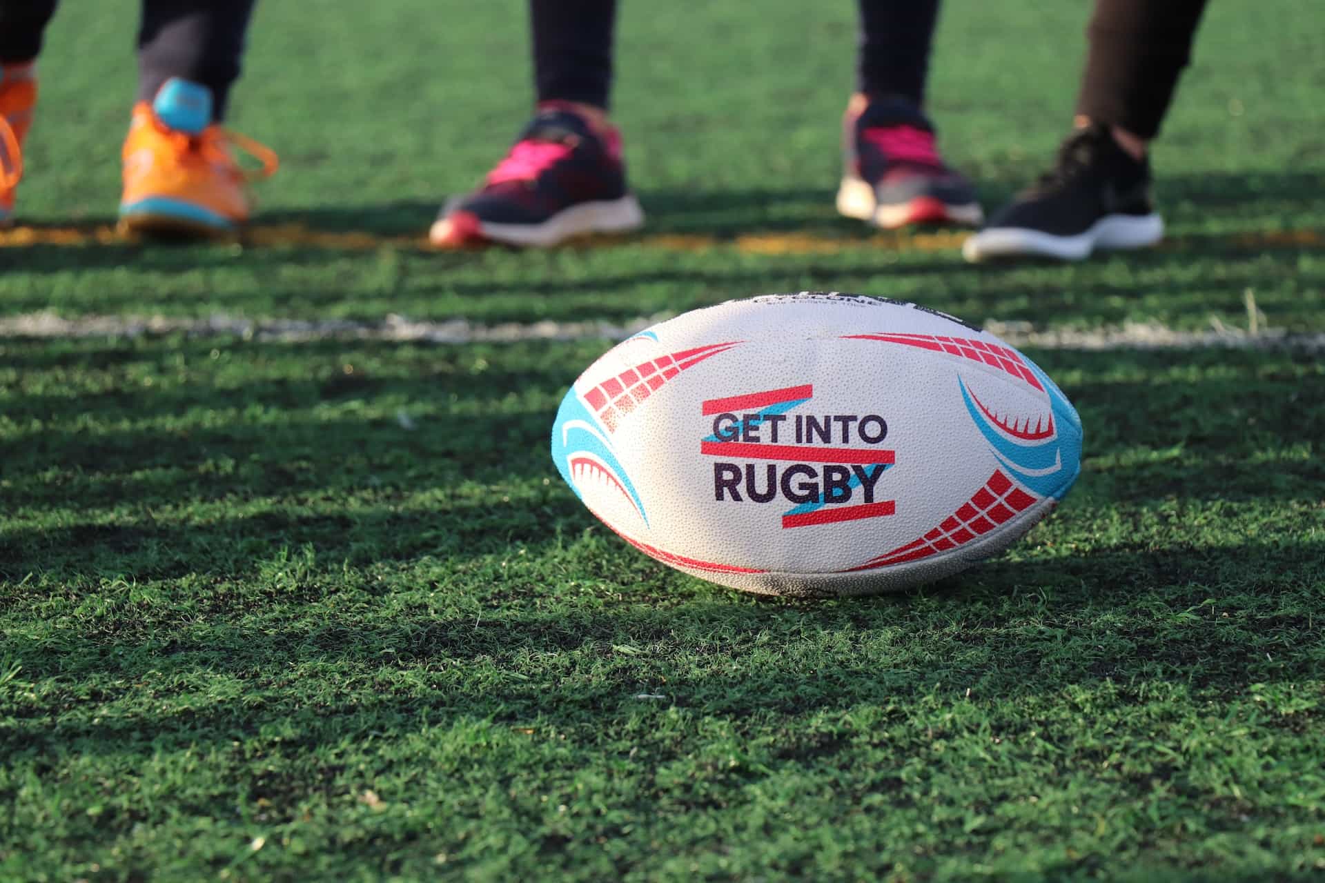 A rugby ball sits on a grass field with people standing in tennis shoes in the background.