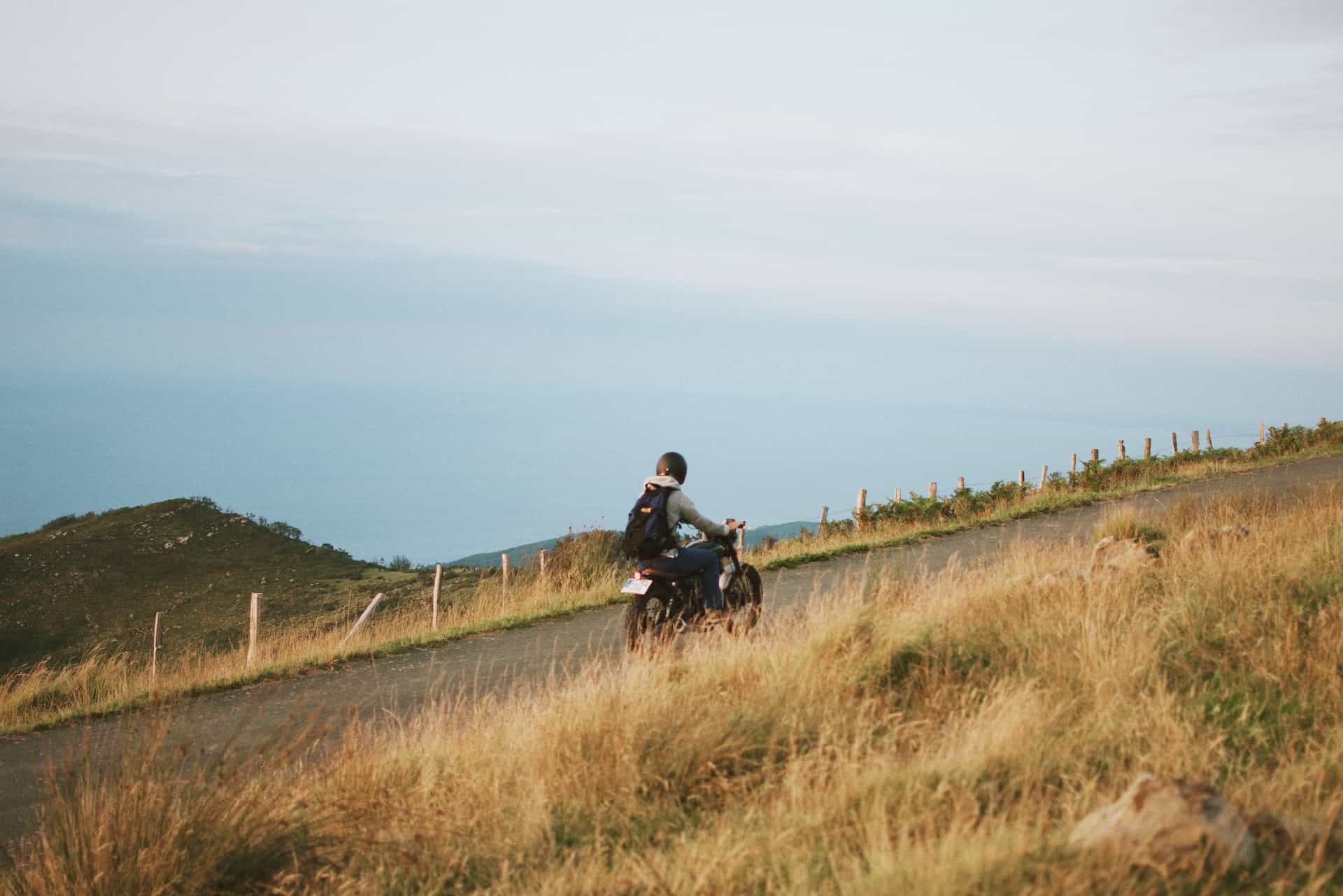 A motorcyclist drives along a small road on a hill, surrounded by fields, overlooking the sea in Basque Country, Spain.