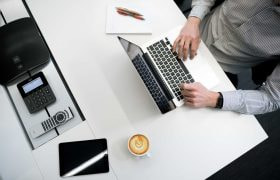 A person using a laptop on a table with a coffee mug, pad, and other gadgets.