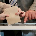Two hands slide a brown envelope into a voting box.