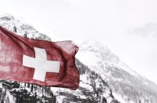 A red flag with a white cross against a snowy mountain range.